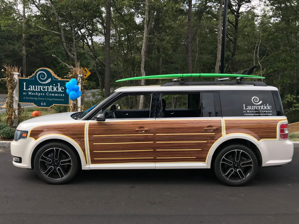 A Ford Flex wrapped in woodgrain and Laurentide at Mashpee Commons branding, parked by the community sign